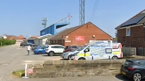 A carpark in front of a small football stadium with a blue covered stand. A sign in the car park says: Welcome to Whitby Town F.C.