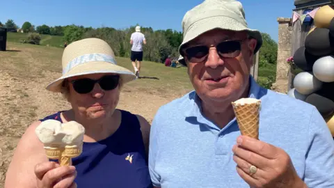 Jeanie and Roger Cole pose together with their ice creams outside. They are an older couple, and are both smiling at the camera and wear hats and sunglasses.