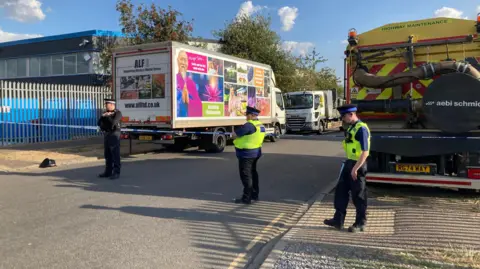 Alex Pope/BBC Three police officers stand alongside a stretch of blue and white police tape, which runs across the road of the industrial estate. There are lorries parked on the road and there is a tall metal fence on the left. 