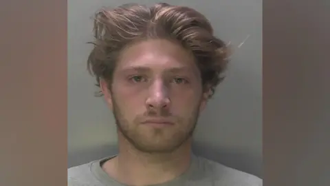 A police mugshot of a man with long hair in a grey top.
