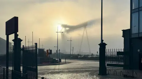 Matthew Poole One of the two Harland and Wolff cranes which dominate the Belfast skyline is obscured by fog on Thursday.   Only a dark outline of the crane is visible because of the thick blanket of fog across the docks area of the city.  The sun is in the background illuminating the crane.