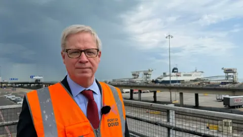 A white haired man with dark framed glasses wearing an orange high viz jacket standing in the port with a ferry in the background