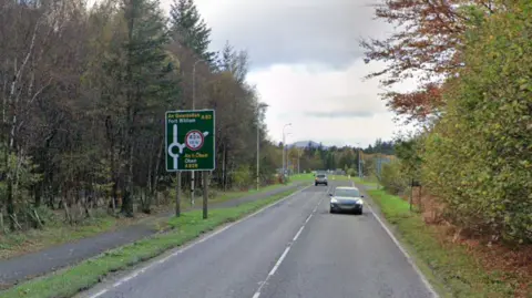 A82 leading up to the Ballachulish roundabout. There are trees at either side of the road and a sign saying a roundabout is coming up. There are two cars on the road.