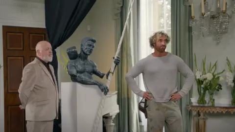 BBC/Chatterbox Media Bill Bailey wears a beige suit and black shirt and stands next to the sculpture, which is holding the long white narwhal tusk. On the other side stands Nick Elphick, who is wearing a grey jumper and jeans with a pattern on them. They are standing in a formal room with curtain swags and fresh flowers behind them.