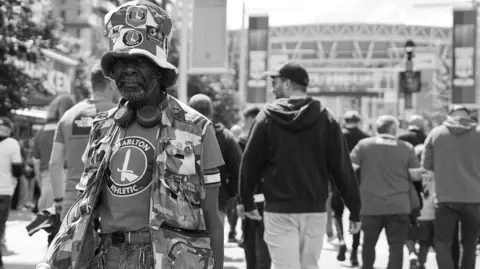 Rhea Spencer-Newell Norman Barker wearing a big Charlton Athletic-themed hat and Charlton t-shirt in front of Wembley Stadium, in a black and white image