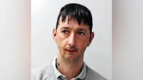 Michael Morrice in a poice mugshot, he is looking slightly to the side of the camera, and is wearing a light-coloured shirt under a grey jumper.