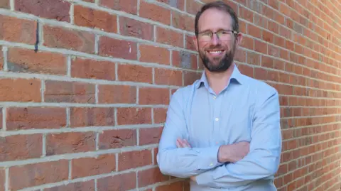 BBC Matthew Barber, Police and Crime Commissioner for Thames Valley, wearing a blue shirt and standing against a brick wall with his arms crossed.