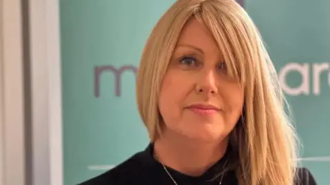 Teresa Cope, a woman with blonde hair and a fringe looks stoically at the camera in front of a Manx Care sign