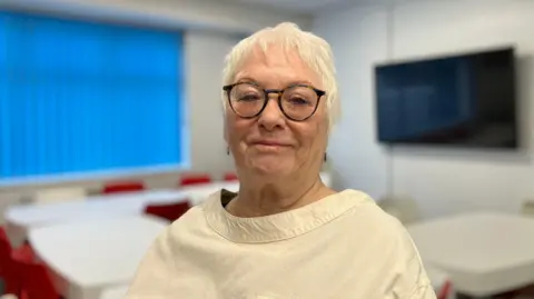 Sam Read/BBC A woman in a beige top looks at the camera. 