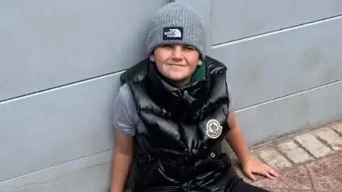 Coby Keogh sits on red brick paving leaning against a grey fence. He is smiling and wearing a shiny black gillet and grey beanie