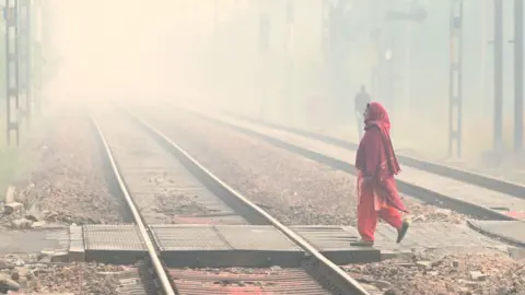 Smog engulfing railway tracks in Delhi on 13 November 2025 in New Delhi, India, as a woman wearing a red Indian outfit, with her head covered walks by. 
