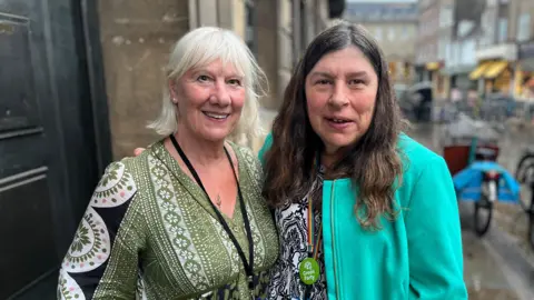 Naomi Bennett, who has long dark hair and is wearing a green jacket over a white and black patterned top, is next to one of her Green party colleagues.