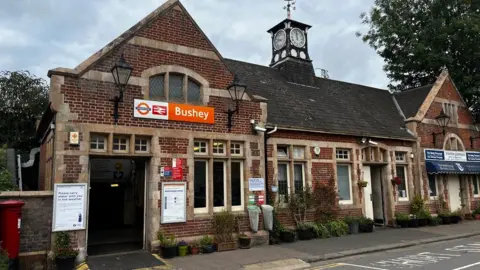 Bryn Holmes/Geograph An old brick-built railway station, with Bushey in white writing on an orange sign to the left. There are posters showing train times to the left and a small shop to the right, with a blue awning.