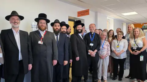 A group of Jewish community leaders and NHS staff standing by the door of the new prayer space while smiling.