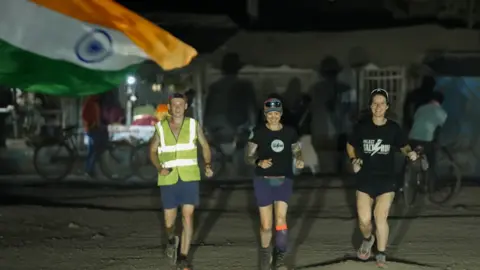 Proyecto Salt Run. Tres corredores, Hannah en el centro, corren en la oscuridad. Una bandera india se ve al fondo.