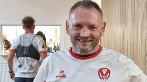 A smiling Gary McKee in a visitor centre wearing a red and white top and inside a community centre where behind him you can see an open hatch 