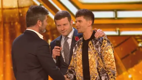 X Factor/ FreemantleMedia/ITV Leon Mallettt on The X Factor stage wearing a golden jacket and shaking hands with Simon Cowell. Dermot O'Leary has his arm around Leon's shoulder.