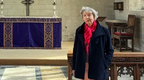 BBC Image of a woman wearing a red scarf and black coat, who is standing in front of an altar in a church.