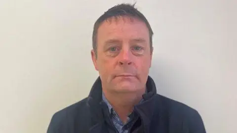A man, Raymond Gormley. Pictured from the shoulders up, he is wearing a blue shirt and a navy jacket. He has brunette hair.