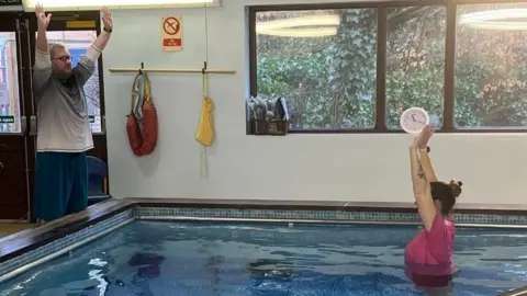 Healthsec Rehab A photograph, supplied by Healthsec Rehab, showing Ball getting physiotherapy at the pool. She is wearing a pink tshirt, and is stood chest-height in the water, side-on to the camera. Her arms are raised above her head and her dark hair is tied back in a bun. On the left of the photo, stood outside the pool, is physiotherapist Jacob Youngson, who is wearing a grey and white long-sleeve top and dark blue shorts. The sides of the pool are dark wood, and the walls are tiled in various shades of blue. There is a large window, looking outside, where trees, ivy, and shrubbery can be seen. 