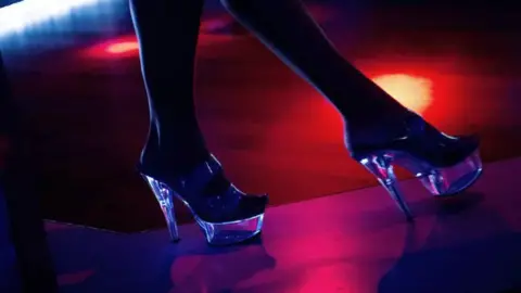 Getty Images A view of someone wearing a pair of clear high heels on stage in a dimly-lit room.