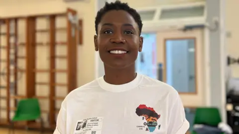 Lesley is wearing a white T-shirt with an Africa logo and a visible visitor badge stands indoors in a hall. Wooden wall bars, chairs, and equipment are visible in the softly blurred background.
