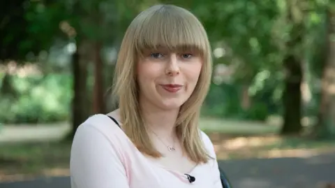 Emily pictured outdoors in a natural setting, wearing a pale pink top against a backdrop of softly blurred greenery.
She has shoulder length blonde hair with a fringe coming down to her blue eyes.