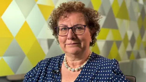 Dame Hilary Cass is sat looking at the camera in a meeting room. She is in front of a glass wall with a geometric pattern.