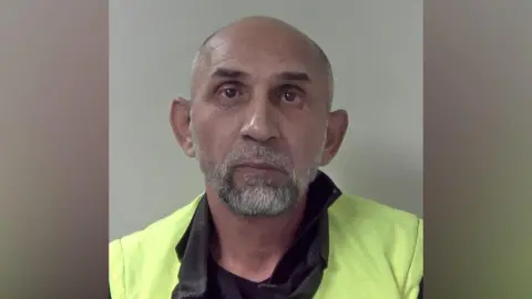 A bald man with a beard wearing a neon yellow jacket in a police mugshot.