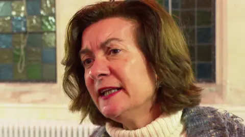 First Minister Eluned Morgan standing in front of stained glass windows in a church-style building. She is wearing a cream coloured polo neck jumper and a multi-coloured cardigan. She has shoulder length wavy brown hair 