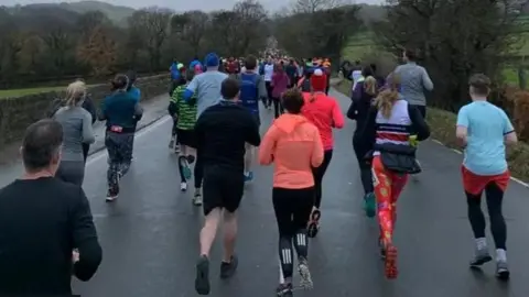Oliver Wright/BBC A group of runners stretching out along a road into the distance. They are mostly dressed in warm clothing on what appears to be a cold Winter day