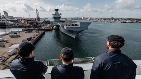 This image captures a scene at a naval port with three individuals in navy uniforms standing on a platform, observing a large aircraft carrier docked nearby. The dock is bustling with vehicles and equipment. In the background, the Spinnaker Tower rises above the buildings.