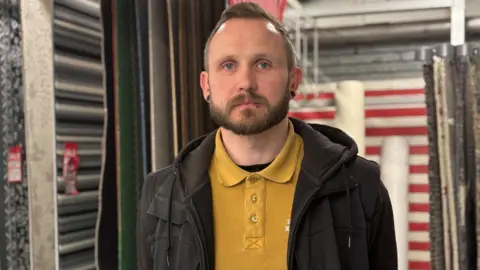 Simon Thake/BBC A young man with closely cropped hair and piercings stands in front of a display of rugs. He is wearing a black hoodie and yellow polo shirt.