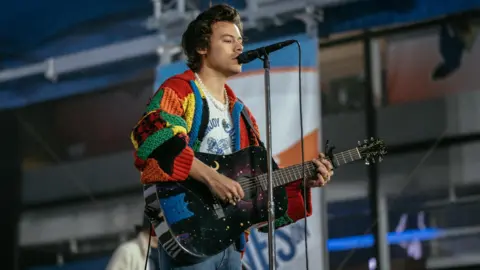 Getty Images Harry Styles stands on a stage holding a black guitar with moon and star details while singing into a microphone. He has longer dark brown hair and is wearing a long pearl necklace, a red, yellow, green and blue patchwork crochet cardigan and a white t-shirt underneath.