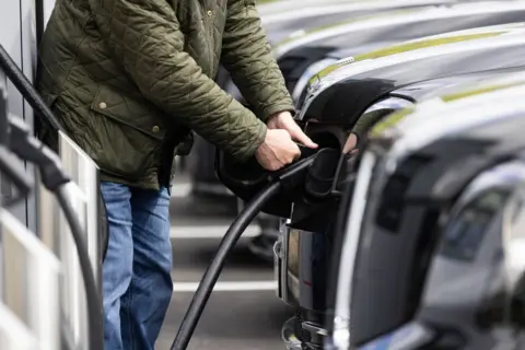 PA Media A close-up of a person fixing a charging cable into a car. They wear blue denim jeans and a green coat.