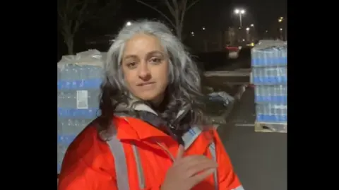 Charlie Maynard A woman wearing an orange jacket stands in a car park at night next to crates of bottled water