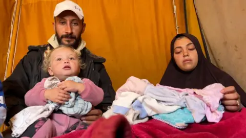 A man holds a child while a woman looks at the child's clothes