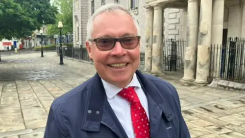 Close up of Councillor Nick Bye. Bye is wearing a white shirt and red tie. He is standing outside and is looking into the camera.