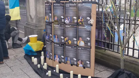 Candles are lying on the ground beside posters of Ukrainian people with details about how the invasion has affected them. The Ukrainian flag is draped over the gates at the city hall.