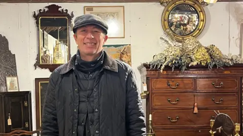 Stuart Warren - a man in a black coat and wearing a green flat cap with a chest of draws and mirrors behind him