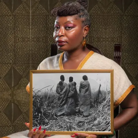Women’s History Museum Zambia Samba Yonga, wearing a beige linen top hemmed with gold-coloured trim, holds up a frame showing an archive photo from the Swedish collection of three women in a field in what is modern-day Zambia, with their backs to the camera, wearing leather cloaks - two children are under the cloaks of two of the women. 
