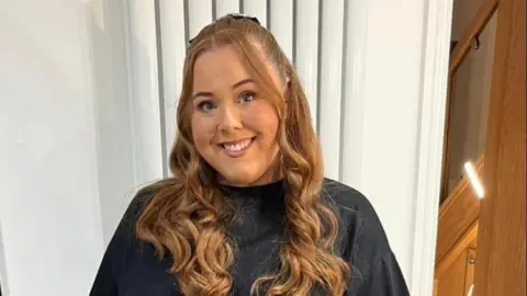 Abbi Hewitson, a woman with long red hair standing and smiling at the camera, wearing a black top. She is smiling. There is a white radiator behind her.