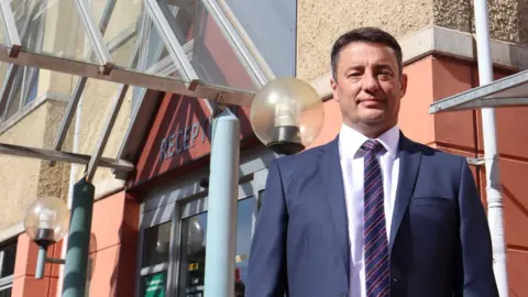 States of Guernsey A man in a blue suit, light pink shirt and striped blue and purple tie stands in front of a modern government building. There is a metal and glass awning above a reception sign.