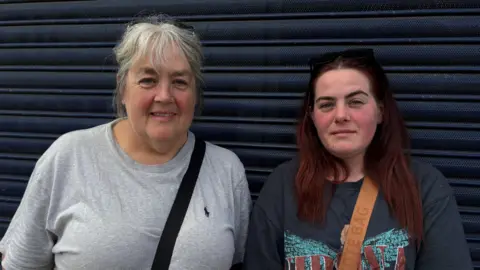 Image shows a lady with a grey t shirt on the left, her grey hair is tied back and has a handbag strap over her shoulder. 
To the right of the picture is a younger lady with a blue Nivarna t shirt on and a brown bag strap. She has red hair.
Both have sun galsses on their head. 
Both are standing in front of blue shutters. 
