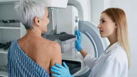 A woman goes for a mammogram.  She is pictured alongside a female medical professional. 