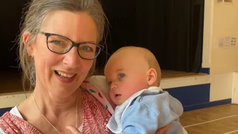 Dorset Council Fru is holding a baby and grinning at the camera. She has greying light brown hair tied back in a low bun and wears black thick-framed glasses and brown and gold stud earrings. She wears and red and white floral print v neck t shirt with a silver chain necklace. The baby has some light blonde hair and blue eyes and wears a pale blue cotton jumper with a white and blue striped bib.