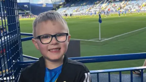 Corby standing in the foreground of a football stadium, close to a blue metal barrier near the pitch. He is wearing dark‑framed glasses, a dark jacket, and a blue top, and is facing the camera with teeth visible. His hair is short and neatly cut, and the face is well lit by daylight.
Behind him, a green football pitch stretches across the image, with a corner flag visible near the touchline. Several players and staff members can be seen in the background wearing training kits. The stadium seating rises behind them, with rows of blue seats and a small number of spectators scattered throughout the stands.
To the right side of the image, another person is walking along the edge of the pitch wearing dark clothing and trainers.