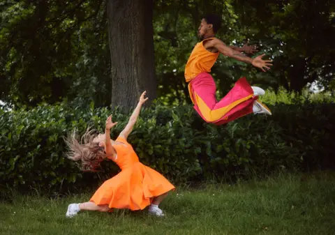 TeesDance Two professional dancers are wearing orange and pink costumes. One has jumped in the air and the other is flipping her hair.