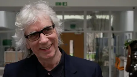Sir Phil Redmond is pictured smiling. He is wearing black-rimmed glasses and has shoulder-length white hair