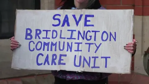 The photo shows someone holding up a white cardboard sign. 'Save Bridlington Community Care Unit' has been written in block capital letters, in an electric blue.